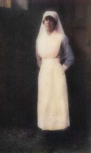 A woman dressed in nurses uniform from the Great War period. Sh has a long sleeved blue dress with a full length white apron, white cuffs and headdress.