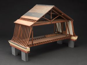 A wooden model of the timber framing of the Mansard roof of a house. It has steep sloping sides and a shallower angled top section.
