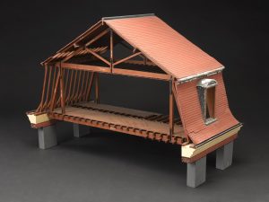 A wooden model of the timber framing of the Mansard roof of a house. It has steep sloping sides and a shallower angled top section.