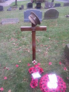 A wooden cross on a grave in Oakworth Cemetery. There are poppy wreaths on the grass in front and an Army cap is perched on the top of the cross.